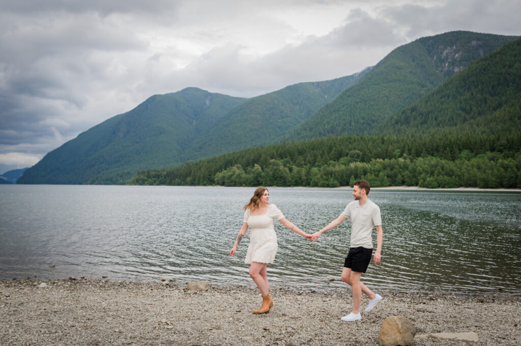 Greenery and mountains in the back ground girl and guy holding hands walking along the Alouette Lake beach.