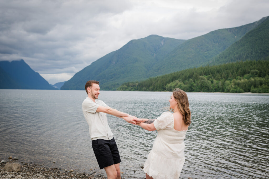 Greenery and mountains in the back ground girl and guy holding hands and spinning in a circle smiling and laughing toward each other.