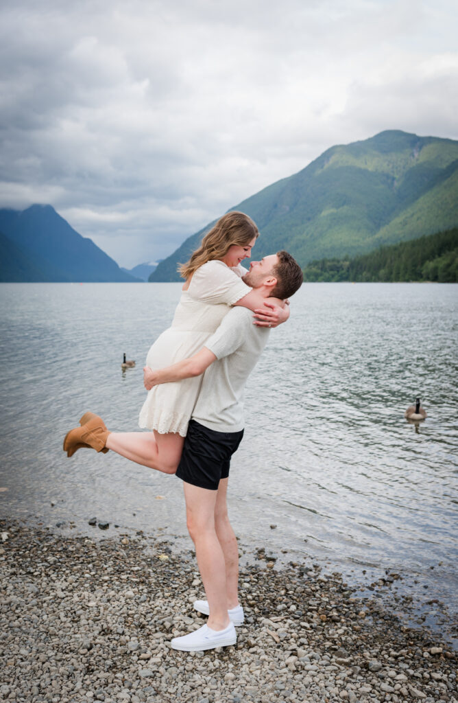 Guy picks up girl for a romantic pose. Girl knees are bent and legs sticking out. Couple looking at each other, lake and mountains in the back round.