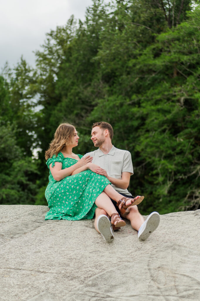 newly engaged couple sitting on a very large rock, girl legs crossed over guys legs. Couple looking at each other.