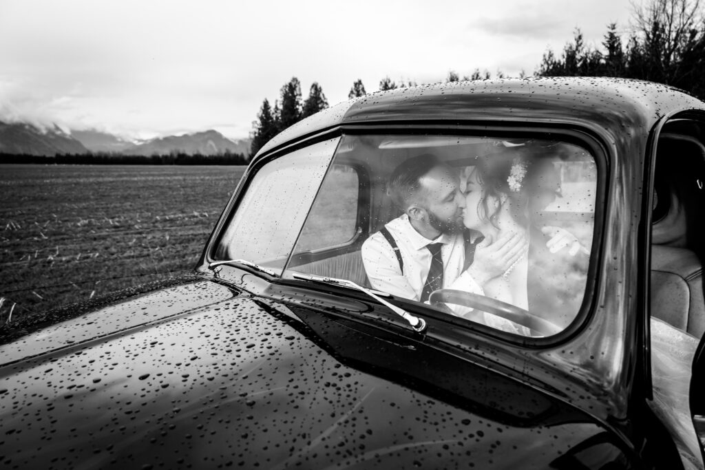 Bride in groom in an old car kissing on their wedding day. Mountains in the back round and rain drops on the car.