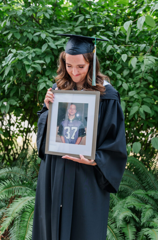 Jessica in her grad outfit holding a frame of her brother and looking down at the frame.  