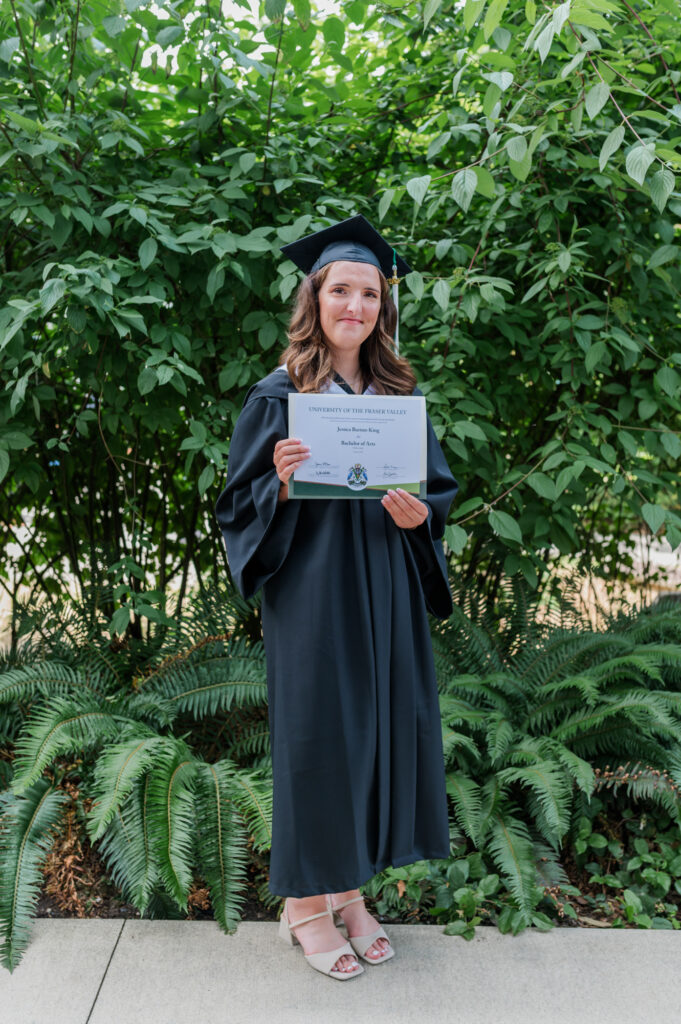 Grad student in there grad and cap gown holding art degree up, greenery in the back round.