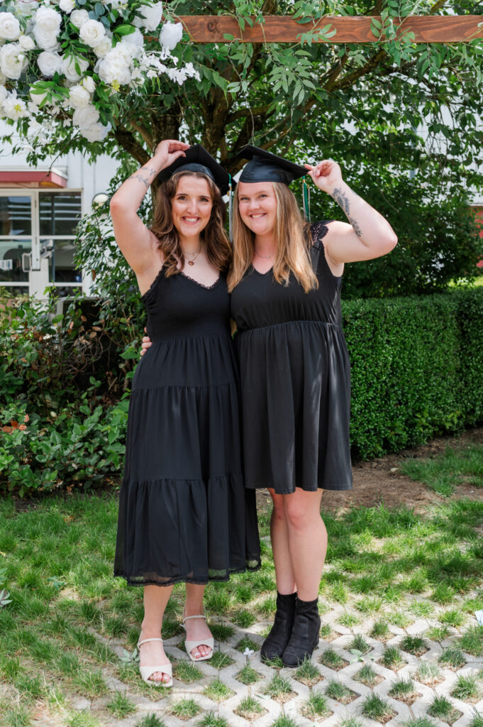 Two grad students holding there cap with one hand and smiling, both wearing black dresses. 