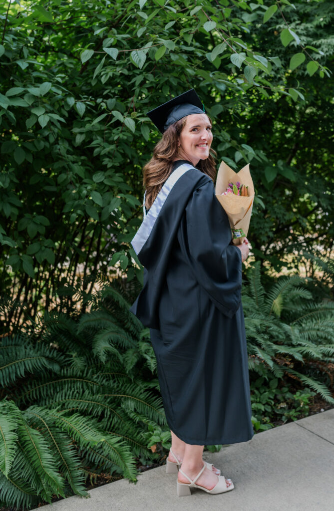 greenery in the back round Grad student in cap & gown holding a bouquet of flowers and looking over her right shoulder.