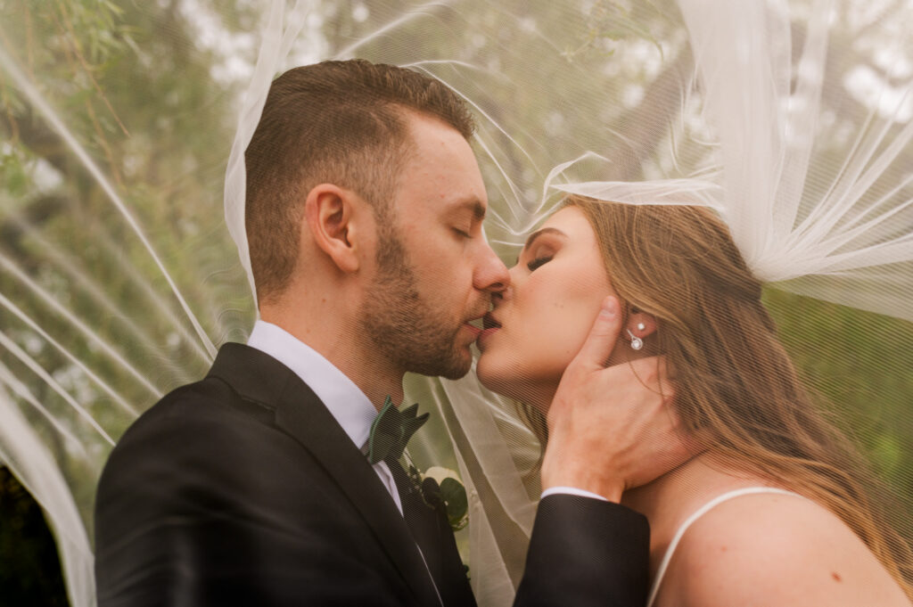 Bride and groom go in for a romantic kiss under the veil. groom holding under brides chin.