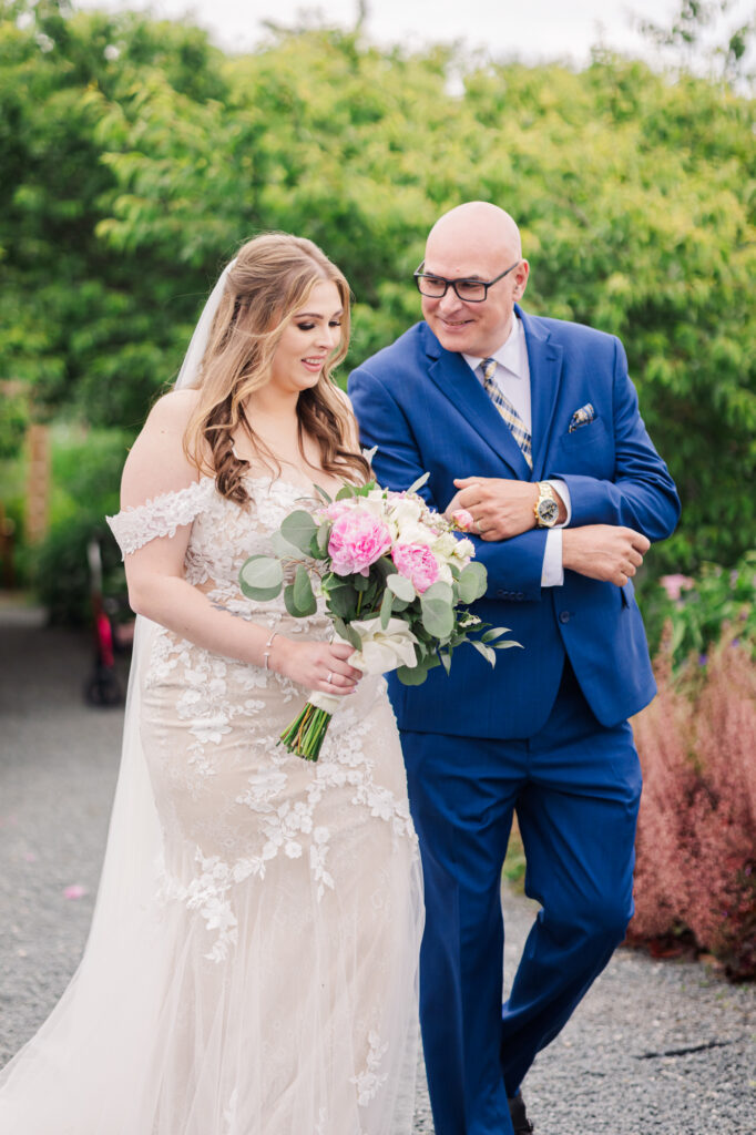 Father of the bride looking at his daughter right before walking down the aisle. Bride looking at bouquet smiling and behind them beautiful garden. 