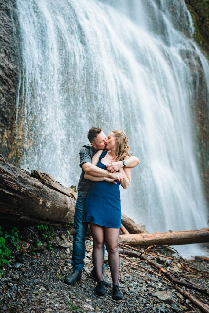 A couple embraces and smiles at each other in a forest setting, surrounded by trees and a rocky riverbed. They appear happy and content.