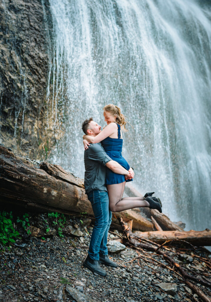 A couple embraces and smiles at each other in a forest setting, surrounded by waterfall, trees and a rocky riverbed. They appear happy and content.