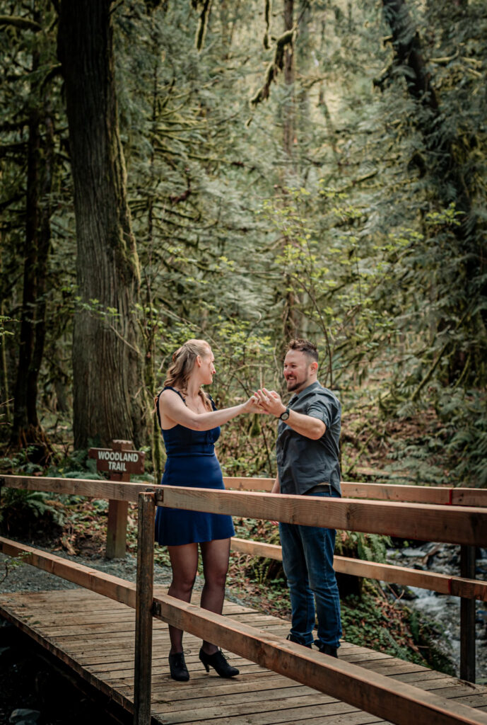 A woman in a blue dress holds hands dancing with a man in a forest by a stream, both smiling. Lush greenery and moss-covered trees surround them.