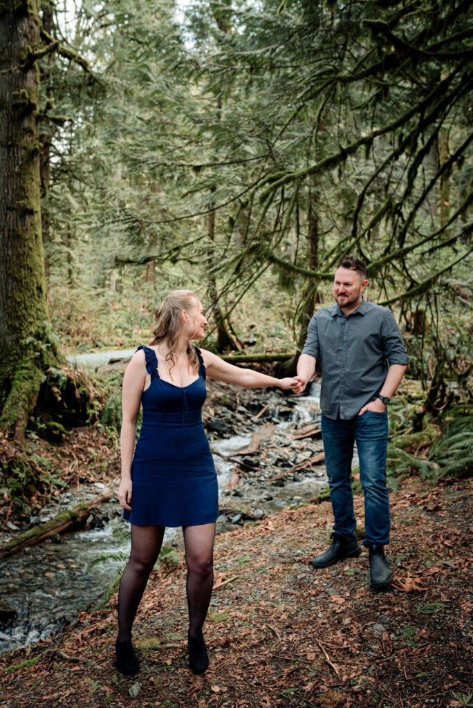 A woman in a blue dress holds hands with a man in a forest by a stream, both smiling. Lush greenery and moss-covered trees surround them.