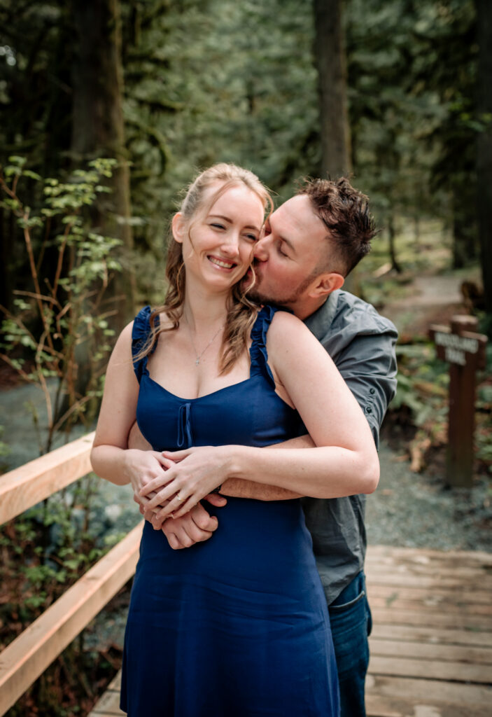 A couple embraces on a wooded path; the man kisses the woman's cheek. She smiles, wearing a blue dress. The setting is lush and green.