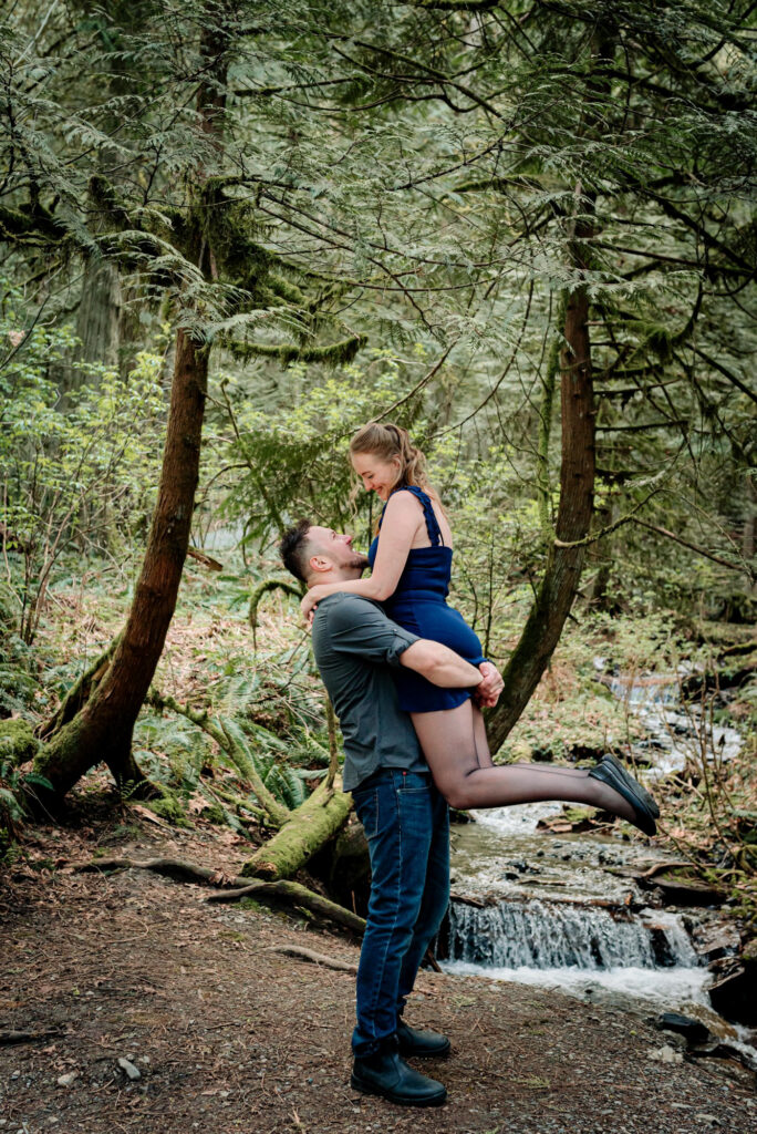 A couple embraces joyfully in a lush forest by a small waterfall. She wears a blue dress, his casual attire. The mood is romantic and serene.