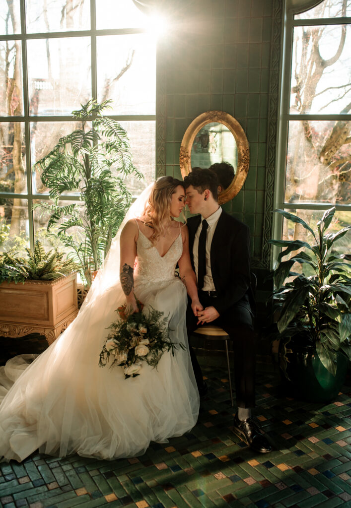 Bride and groom lean in for a kiss, sitting on a chair and holding one hand with green plants surrounding them. 