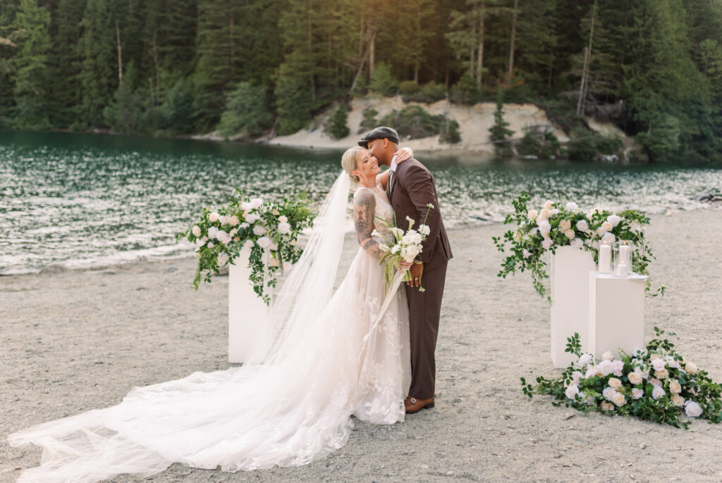 Groom whispering into brides ear and bride in white holding her arm on grooms shoulder. Beach back round and flower bouquet around them. 