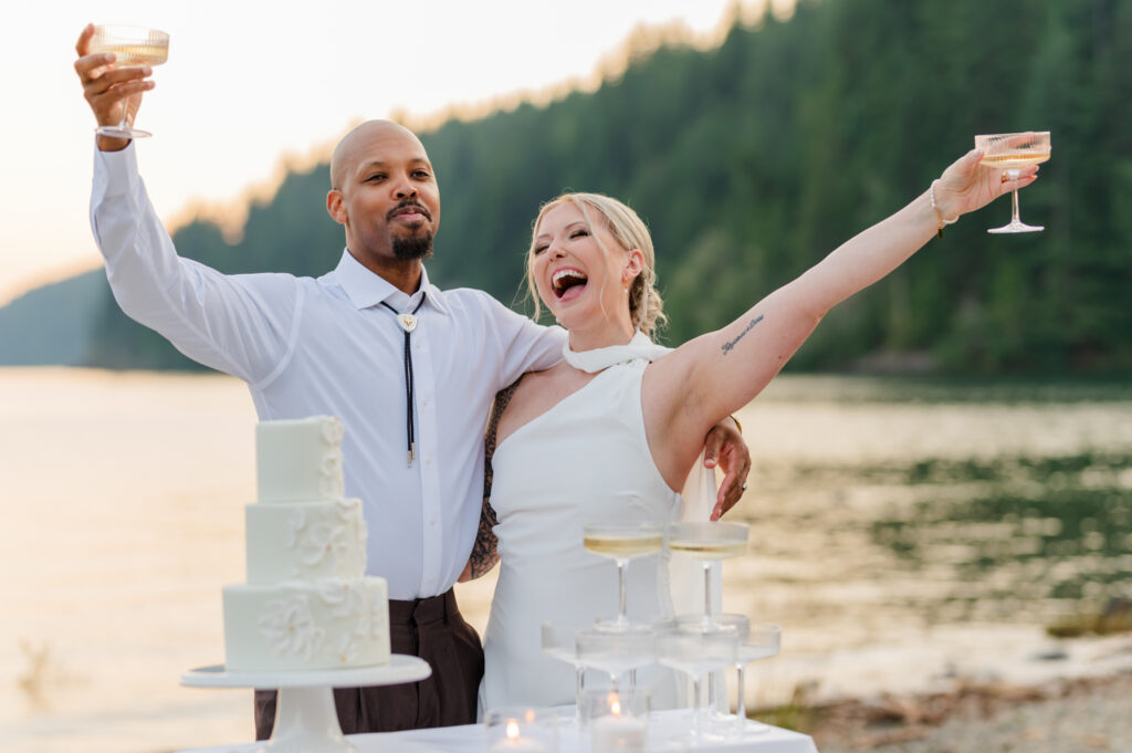 Bride and groom holding their champagne glasses up and so excited with laughter, Cake and beach in the back area.