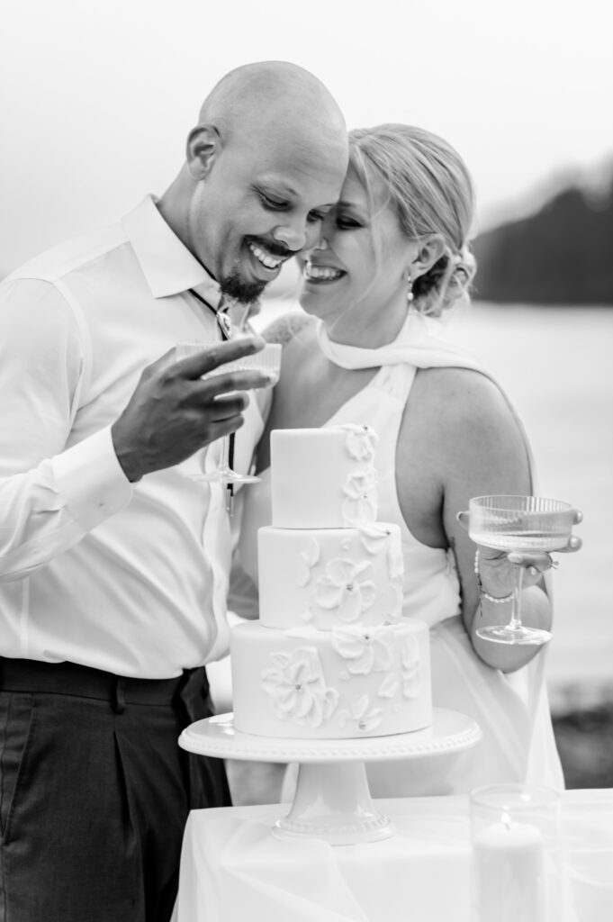 Black and white photo of bride and groom dressed in their wedding attire. Both holding champagne glasses in one hand and smiling very close to each others foreheads. 