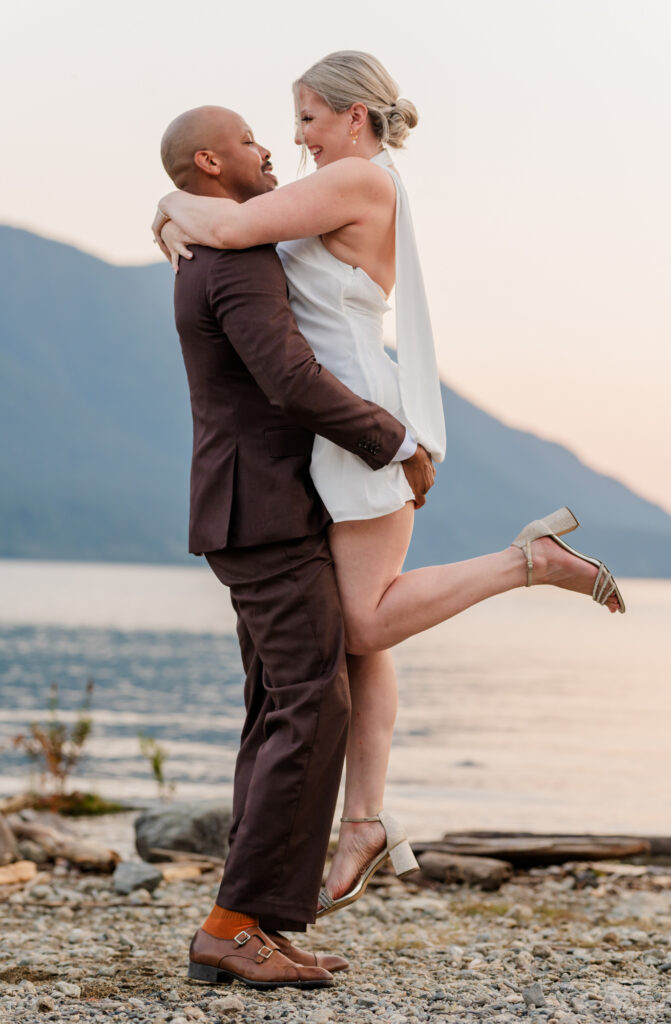 Groom in brown suite and Bride in white summer dress, groom lifts the bride up and bride lifts one leg up. Holding the grooms shoulders looking at each other madly in love. 