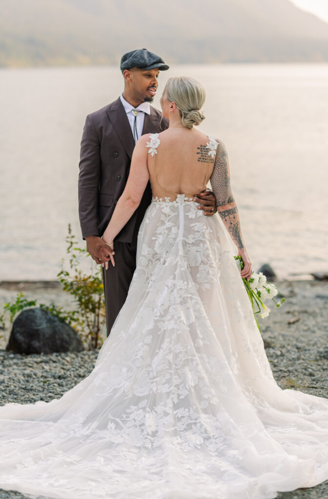 Bride and groom looking at each other , groom holding brides hand. You can see the brides dress from behind her back. Beach and mountains in the back round. 
