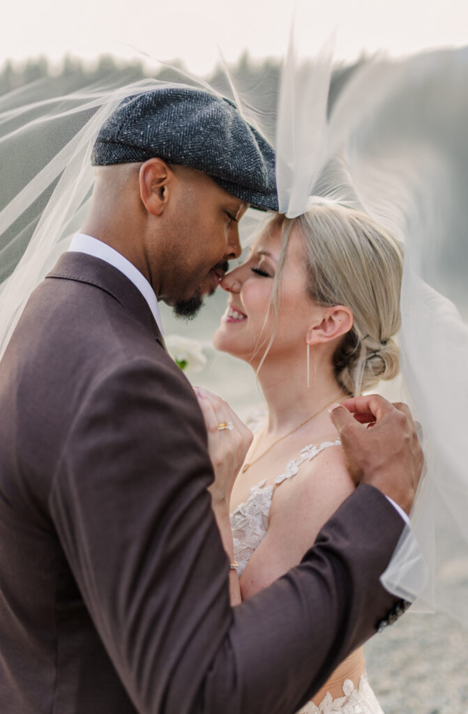 Bride and groom touching noses both eyes are closed, they are under the brides wedding veil dressed in their wedding attire. 