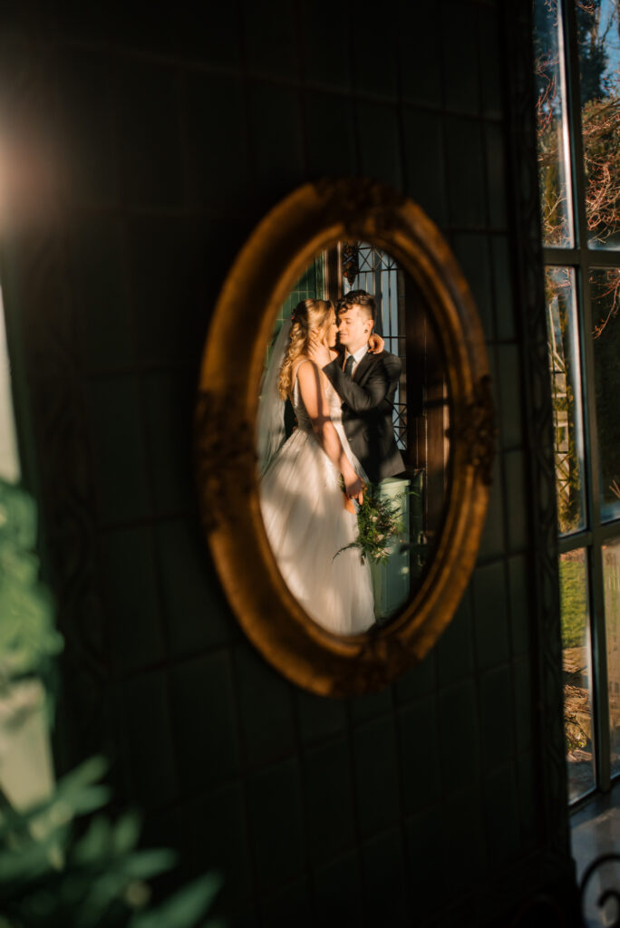 Bride and groom snuggle in for a kiss but first touching each other nose. Photo taking them in a mirror. 