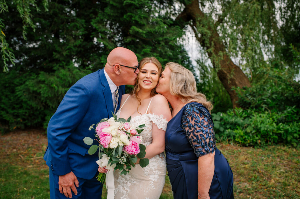 Mom and dad kissing daughters cheek on her wedding day, bride smiling big and holding flower bouquet.