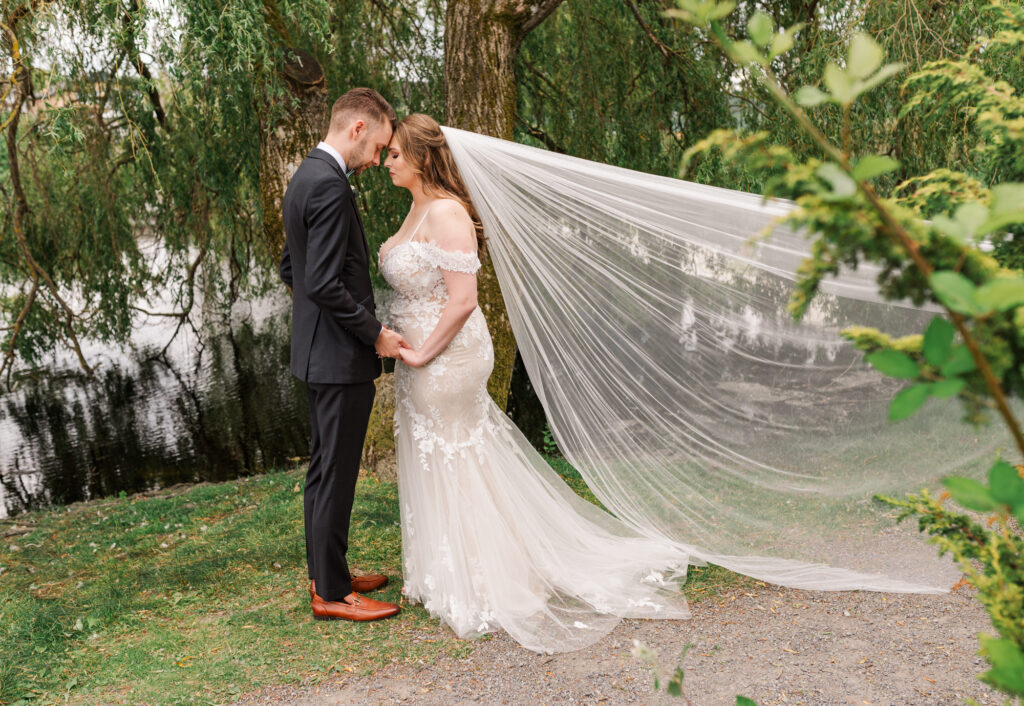 Bride and groom foreheads touching, holding hands and willow trree in the back round.