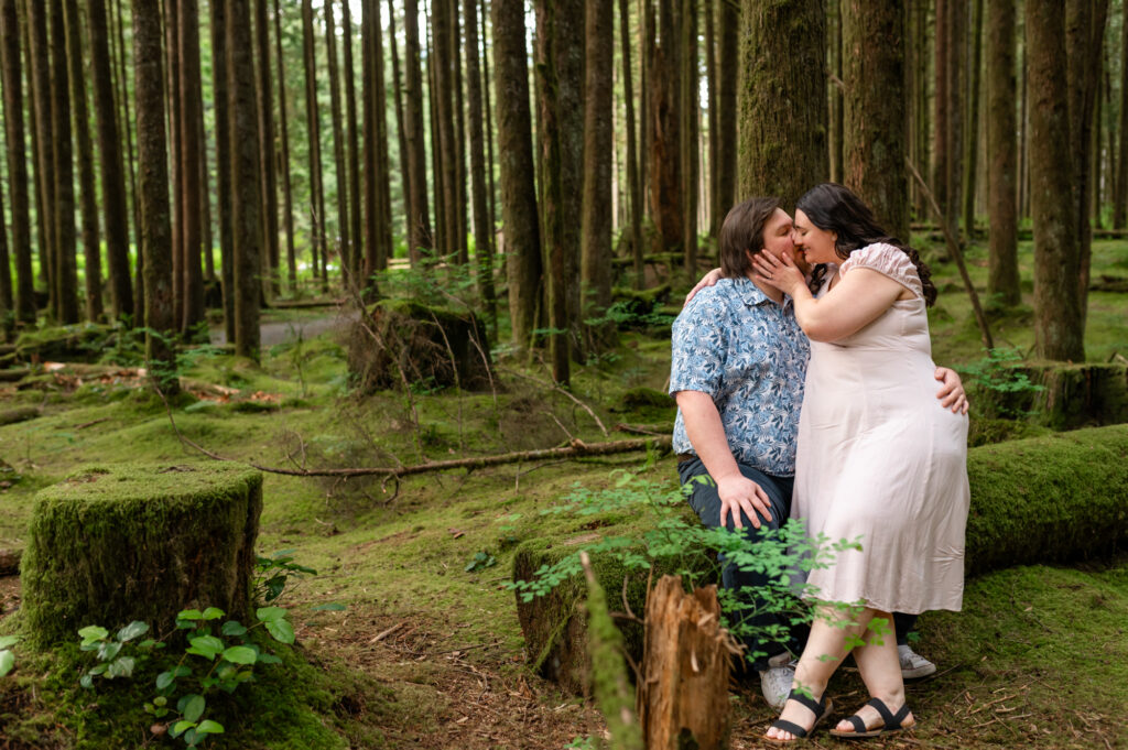 Golden Ears Engagement couple sitting and kissing, greenery and tall trees in the back round.